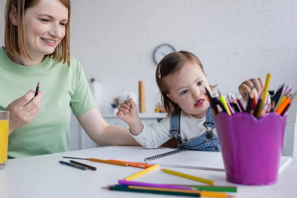 Criança com síndrome de down tomando lápis de cor perto de mãe sorridente e suco de laranja na cozinha — Fotografia de Stock