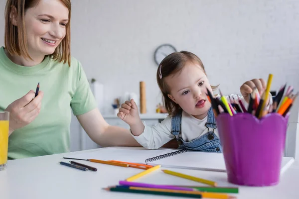 Enfant atteint du syndrome du duvet prenant crayon de couleur près de maman souriante et du jus d'orange dans la cuisine — Photo de stock