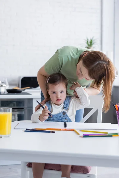 Woman hugging daughter with down syndrome near color pencils and orange juice in kitchen — Stock Photo