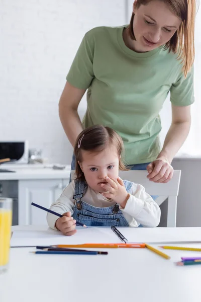 Niño con síndrome de Down sosteniendo lápiz de color cerca de la madre y cuaderno de bocetos en la cocina - foto de stock