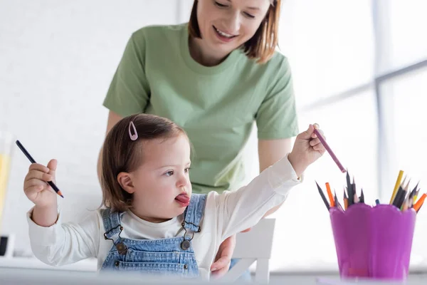 Niño con síndrome de Down tomando lápiz de color cerca de mamá sonriente en casa - foto de stock