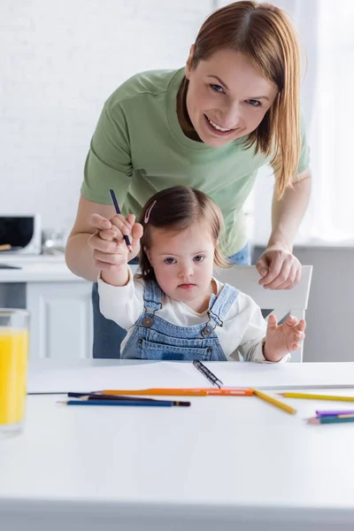 Donna sorridente guardando la fotocamera vicino al bambino con sindrome di Down e matite di colore — Foto stock