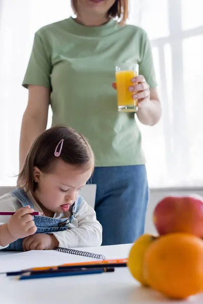 Niño con síndrome de Down sosteniendo lápiz de color cerca de papel y mamá borrosa con jugo de naranja - foto de stock
