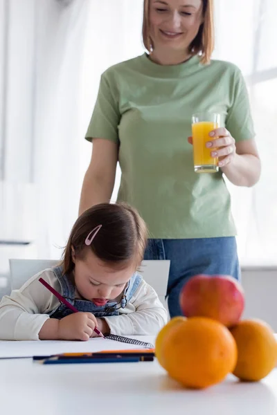 Kid with down syndrome drawing near smiling mom with orange juice in kitchen — Stock Photo