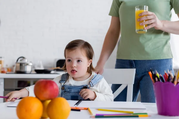 Menina da criança com síndrome de down segurando lápis de cor perto de frutas e mãe com suco de laranja na cozinha — Fotografia de Stock