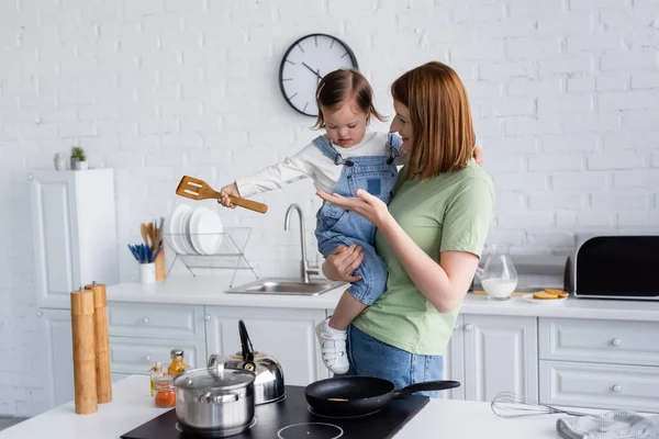 Smiling woman holding daughter with down syndrome holding spatula near stove in kitchen — Stock Photo