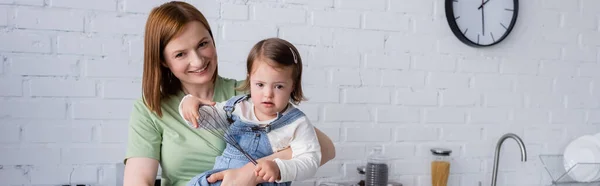 Donna sorridente che tiene la figlia con la sindrome di Down che tiene frusta in cucina, banner — Foto stock