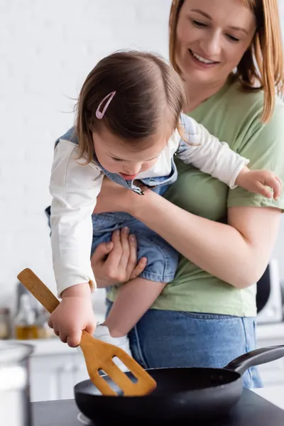 Mujer sonriente sosteniendo niño con síndrome de Down mientras cocina en la cocina - foto de stock