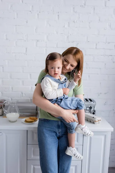 Feliz madre sosteniendo hija con síndrome de Down y batir en la cocina - foto de stock