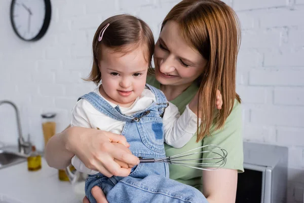 Kid with down syndrome holding whisk near smiling mom in kitchen — Stock Photo