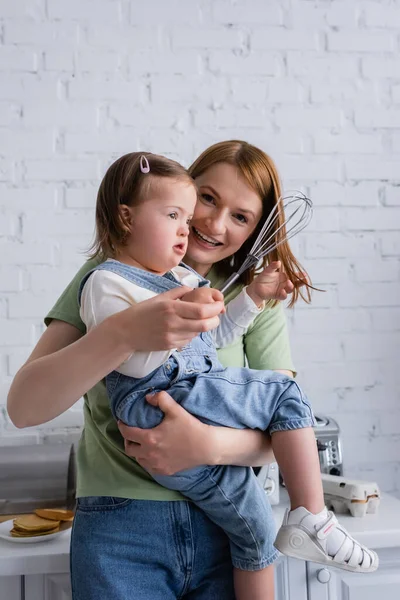 Un parent positif tenant un enfant atteint du syndrome du duvet tenant un fouet dans la cuisine — Photo de stock