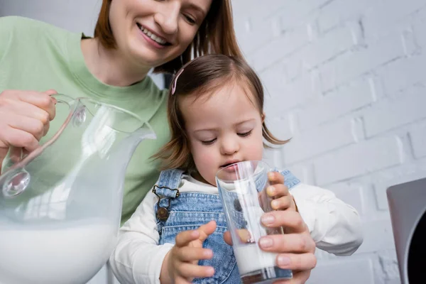 Mujer sonriente sosteniendo jarra con leche cerca de la hija con síndrome de Down en la cocina - foto de stock