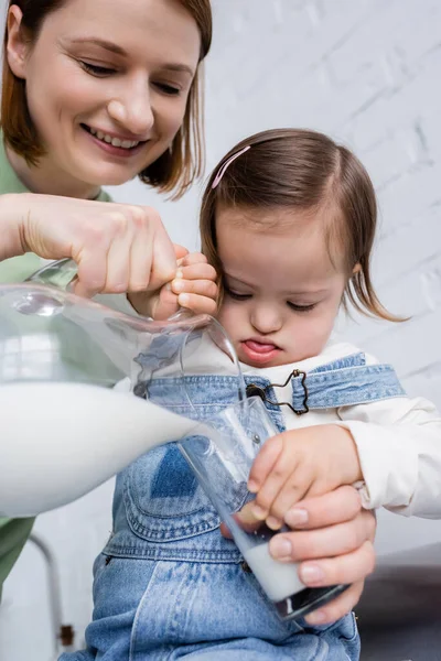 Smiling parent pouring milk from jug near kid with down syndrome in kitchen — Stock Photo