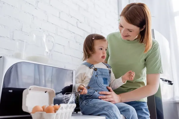 Sonriente madre sosteniendo hija con síndrome de Down cerca de los huevos y la leche en la cocina - foto de stock