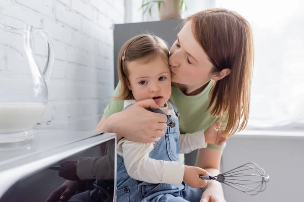 Woman kissing daughter with down syndrome holding whisk in kitchen — Stock Photo