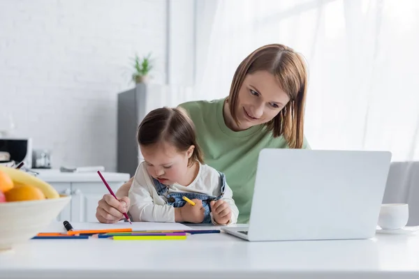 Sonriente freelancer mirando portátil cerca de hija con síndrome de Down dibujo en la cocina — Stock Photo