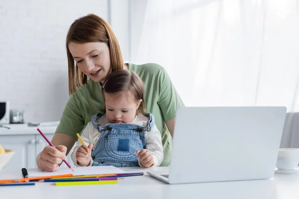 Donna felice disegno vicino bambino con sindrome di Down vicino laptop e caffè in cucina — Foto stock