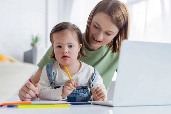 Woman drawing near daughter with down syndrome and blurred laptop in kitchen — Stock Photo