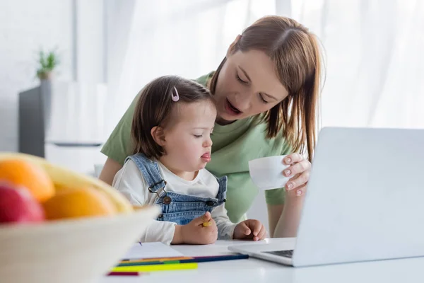 Woman holding cup near laptop and daughter with down syndrome holding color pencil — Stock Photo