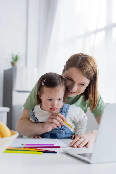Mujer positiva sosteniendo lápiz de color cerca del ordenador portátil y la hija con síndrome de Down en la cocina - foto de stock