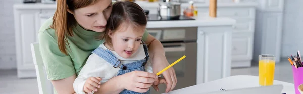 Mamá sosteniendo lápiz de color cerca del niño con síndrome de Down y jugo de naranja en la cocina, pancarta - foto de stock