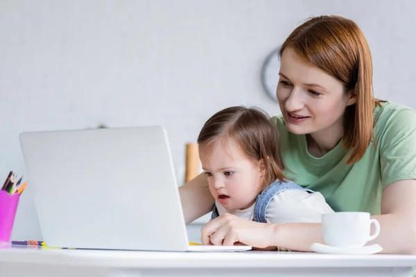 Freelancer sonriente usando laptop cerca de su hija con síndrome de Down y café en casa — Stock Photo