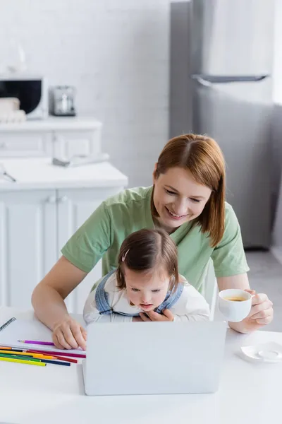 Madre e hijo con síndrome de Down mirando el portátil cerca de lápices de color en la cocina - foto de stock