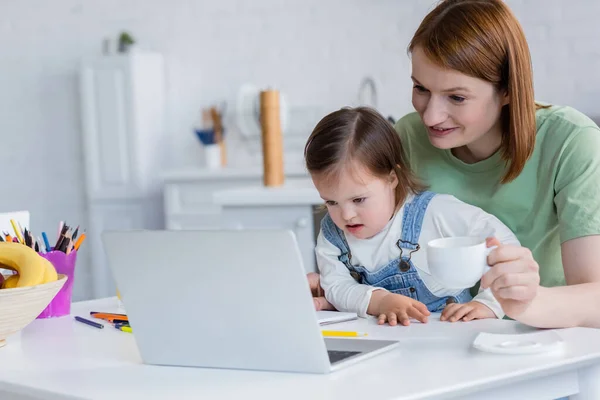 Femme souriante tenant tasse près de l'enfant avec le syndrome du duvet, ordinateur portable et crayons de couleur dans la cuisine — Photo de stock