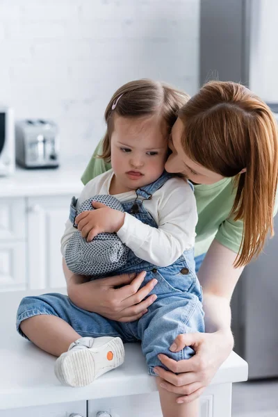 Madre abbracciando figlia con sindrome di Down attaccare fuori la lingua e tenendo guanto da forno in cucina — Foto stock
