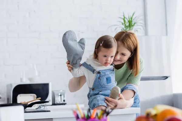 Улыбающаяся женщина стоит рядом с ребенком с синдромом Дауна и выпечкой перчатки на кухне — стоковое фото