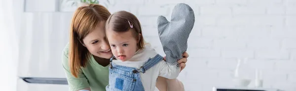 Madre feliz mirando al niño con síndrome de Down y guante para hornear en la cocina, pancarta - foto de stock