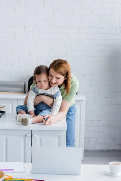 Femme souriante pointant vers un ordinateur portable près de la fille avec le syndrome du duvet dans la cuisine — Photo de stock