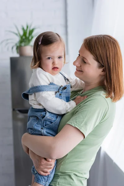 Mulher sorridente olhando para a filha do bebê com síndrome de down na cozinha — Fotografia de Stock