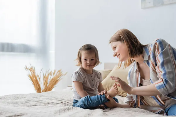 Улыбающаяся мать касается ног маленькой дочери с синдромом Дауна на кровати — стоковое фото