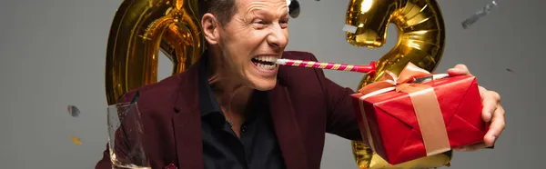 Homme excité avec corne de fête, champagne et cadeau près des ballons sur fond gris, bannière — Photo de stock