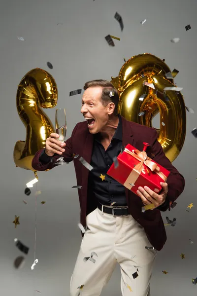 Homme mûr excité avec présent et verre de champagne près de ballons et confettis sur fond gris — Photo de stock