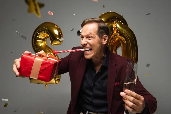 Aufgeregter Mann mit Partyhorn hält Geschenkbox und Champagner in der Nähe von Luftballons und Konfetti auf grauem Hintergrund — Stockfoto