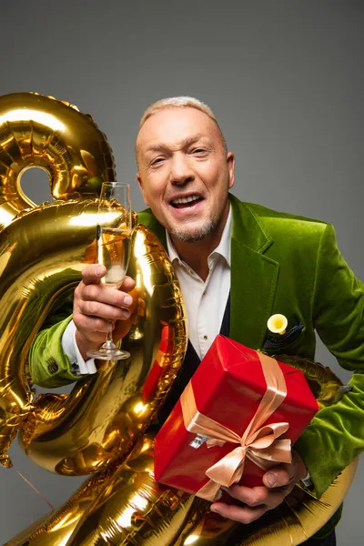 Homme mûr souriant avec présent et champagne regardant la caméra près de ballons isolés sur gris — Photo de stock