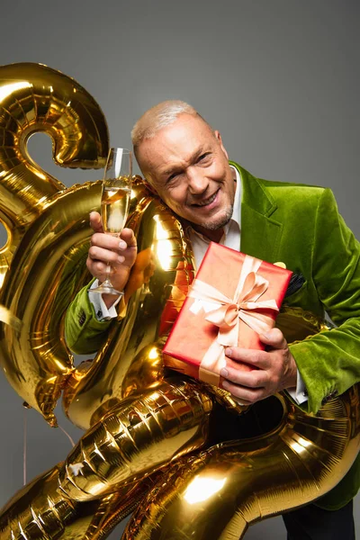 Mature man holding gift and glass of champagne near balloons isolated on grey — Stock Photo