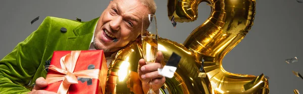 Middle aged man with champagne and present looking at camera near balloons and confetti on grey background, banner — Stock Photo