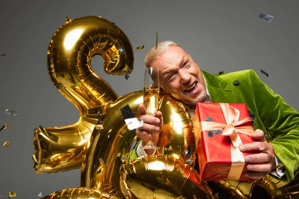 Positive reife Mann hält Champagner und Geschenk in der Nähe von Konfetti und Luftballons auf grauem Hintergrund — Stockfoto