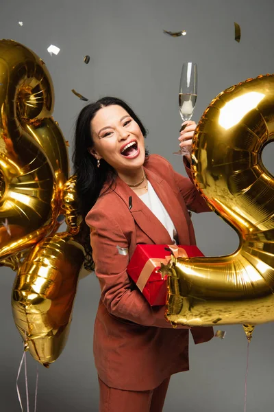 Femme asiatique excitée tenant un verre de champagne et présent près de confettis et ballons sur fond gris — Photo de stock