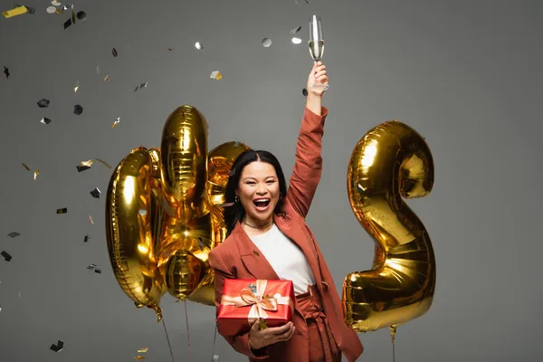 Захоплена азіатська жінка тримає подарунок і шампанське біля кульок і конфетті на сірому фоні — стокове фото