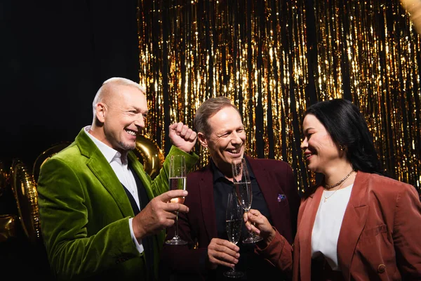 Cheerful interracial friends holding glasses of champagne near tinsel on black background — Stock Photo