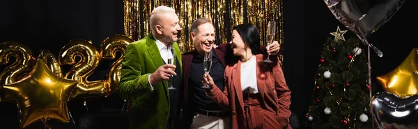 Amis interraciaux positifs tenant des verres de champagne près de ballons, arbre de Noël sur fond noir, bannière — Photo de stock