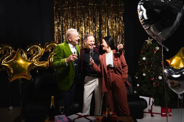 Amigos multiétnicos positivos con champán cerca de globos y árbol de Navidad sobre fondo negro - foto de stock