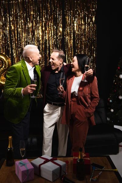 Excited interracial people with champagne talking near gifts near tinsel on black background — Stock Photo