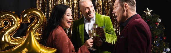 Excited multiethnic friends with champagne talking near tinsel and balloons on black background, banner — Stock Photo