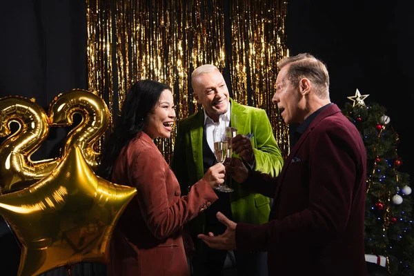 Homme excité tenant du champagne et regardant des amis interraciaux près de l'arbre de Noël et des ballons sur fond noir — Photo de stock