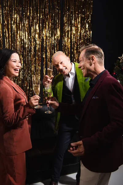 Happy multiethnic mature people holding glasses of champagne near tinsel on black background — Stock Photo